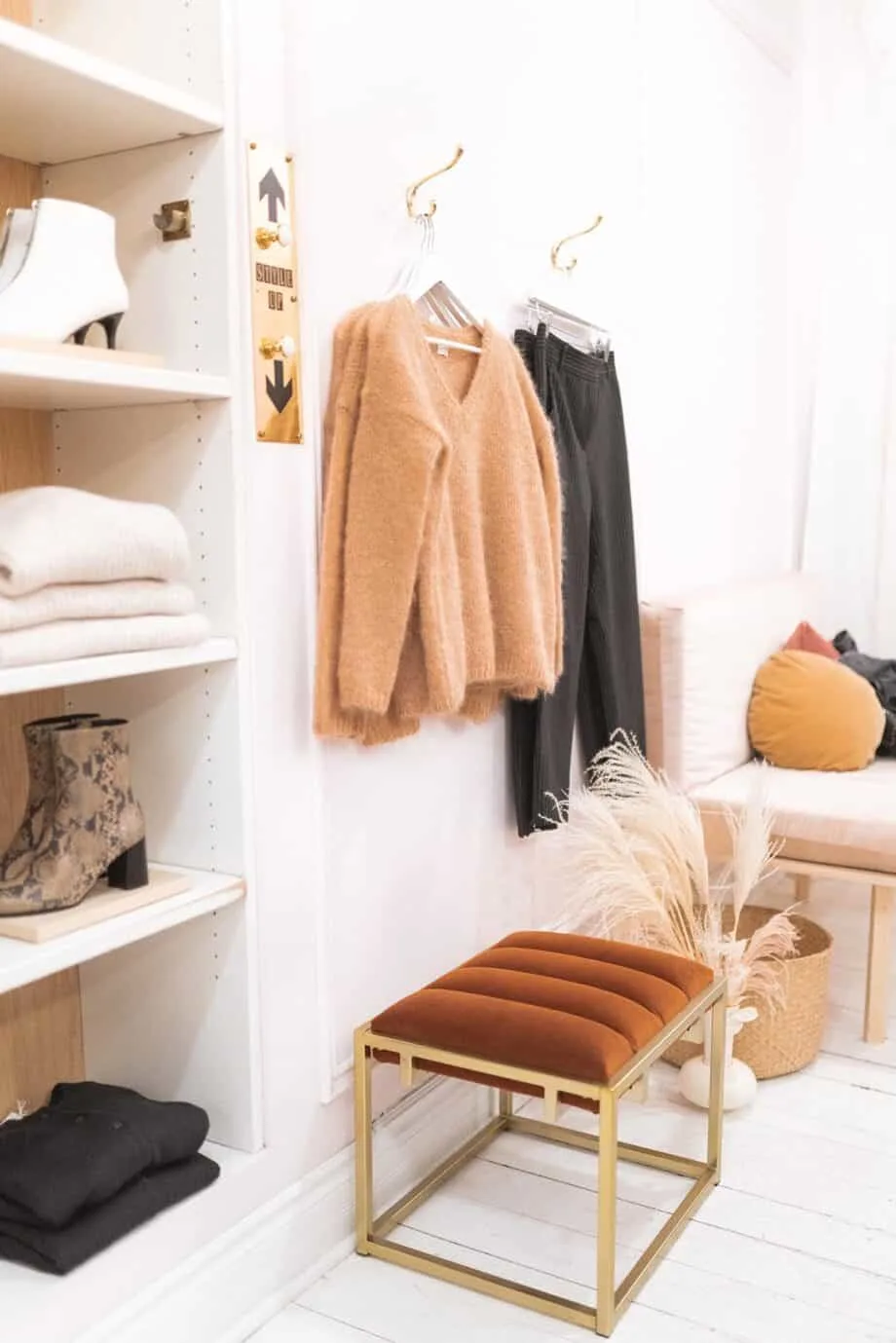 Inside of a wardrobe with a stool and shelves showing clothes folding, as well as a cardigan and pants hanging on hooks